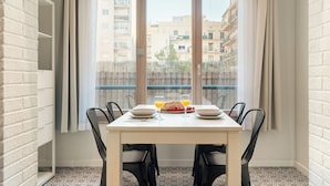 A dining area with a table set for two, a window with curtains, and a view of buildings outside. - Fuster Apartments By Aspasios  (Barcelona)