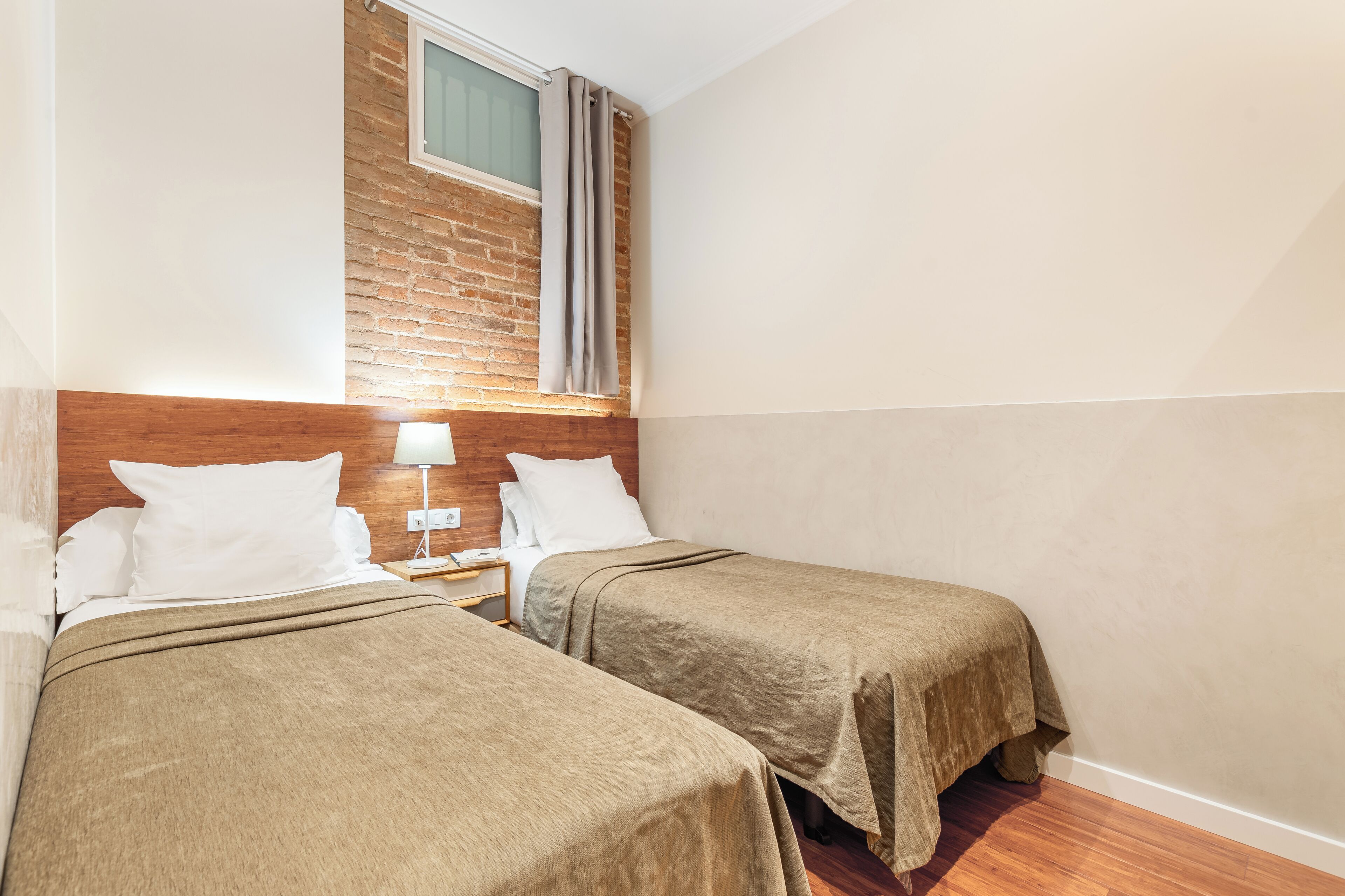 Habitación de hotel con dos camas, un cabecero de madera, una mesita de noche con lámpara y una pared de ladrillo.