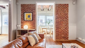 A modern living room with a brick wall, a leather sofa, a dining area with a table and chairs, and a view through an open door. - Fuster Apartments By Aspasios  (Barcelona)