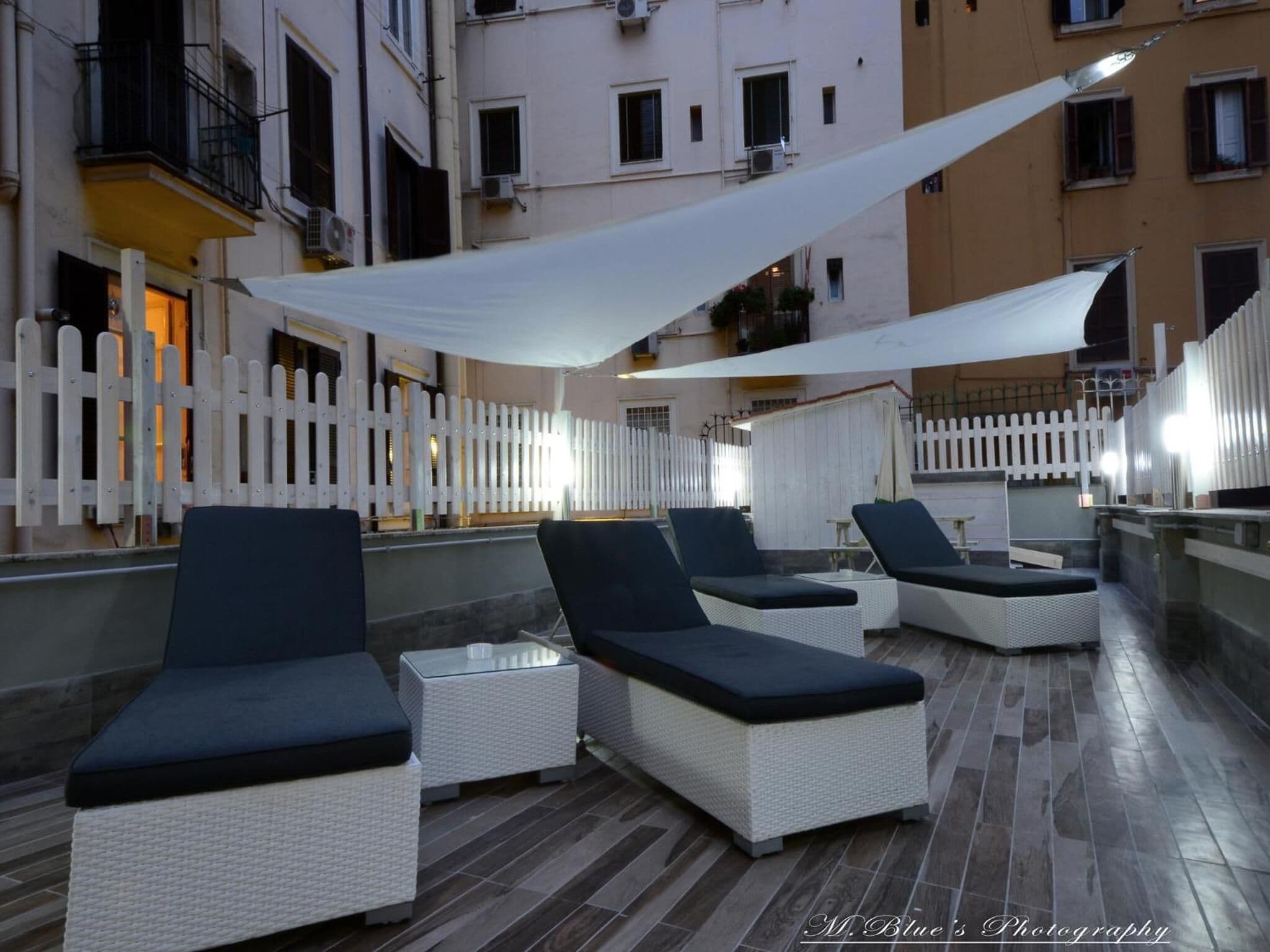 An outdoor seating area with lounge chairs and tables, a white fence, and buildings in the background.