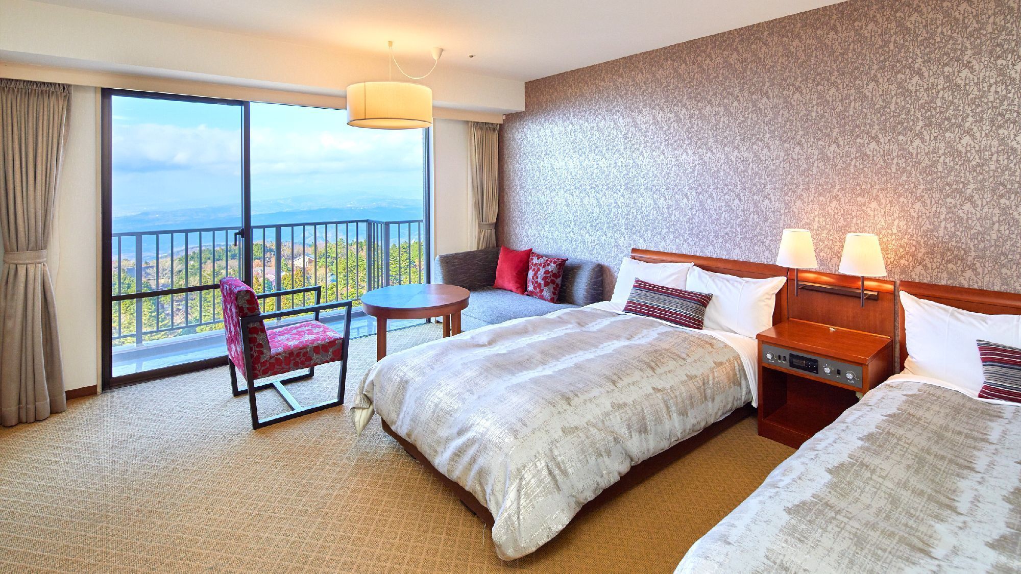 A hotel room with a large bed, a sofa, a chair, a small table, and a nightstand. There is a view of the outdoors through a large window.