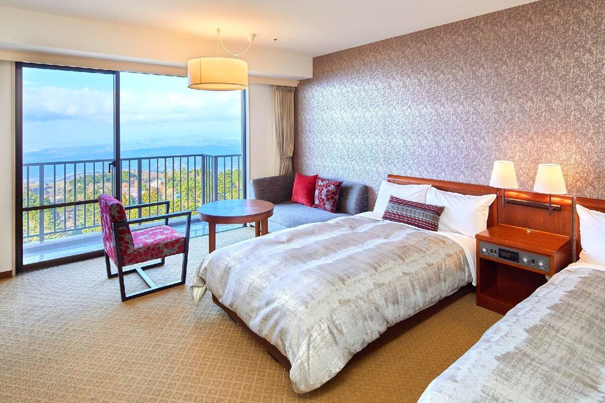 Habitación de hotel con una cama grande, un sofá, una silla, una mesita y una mesita de noche. Se aprecia vista al exterior a través de un ventanal amplio.