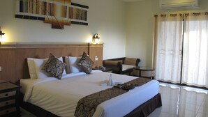 Quarto de hotel com uma cama grande, mesa de cabeceira, cadeira, obra de arte na parede, ar condicionado e janela com cortinas.