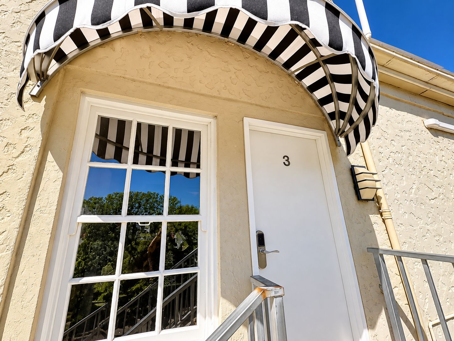A building entrance with a white door marked with the number 3, a window with a striped awning, and a metal handrail.