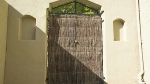 Une grande porte en bois fermée, surmontée d’une barre métallique, adossée à un mur sobre agrémenté de deux petites fenêtres en arc de cercle.