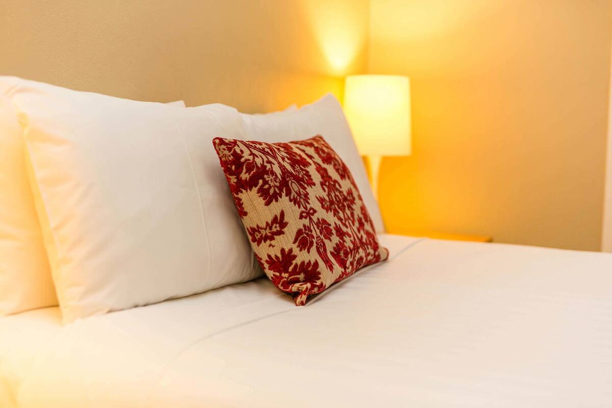 A bed with white bedding and a single decorative pillow with a red floral pattern.