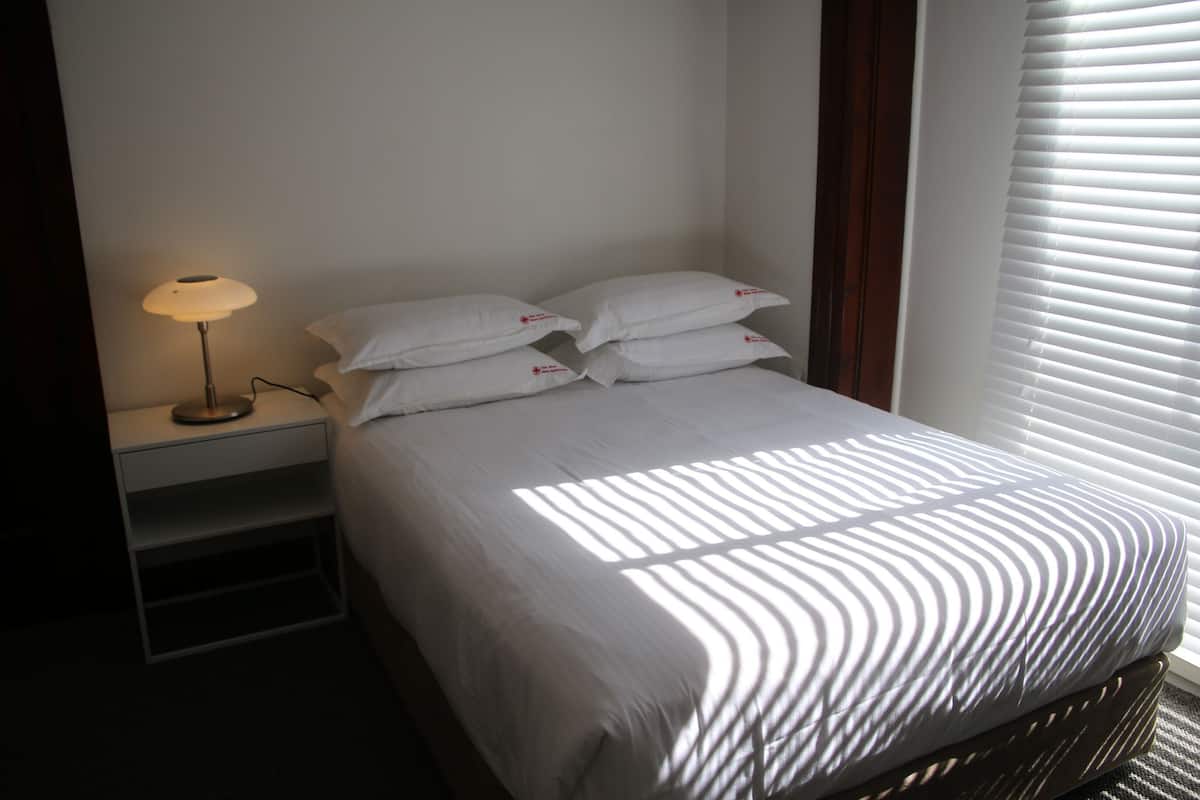 A neatly made bed with white linens and pillows, a bedside lamp, and a window with blinds.