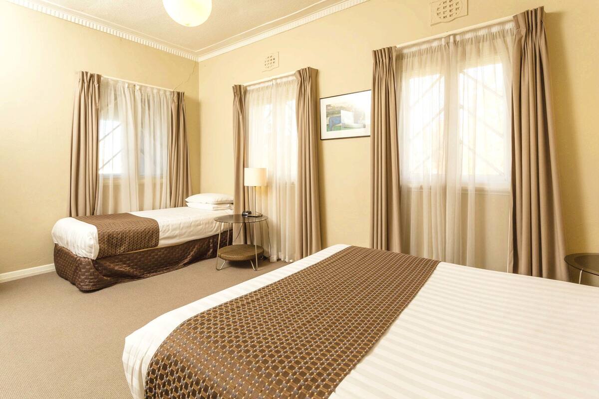 A hotel room with two beds, a small table, a lamp, and a window with curtains.