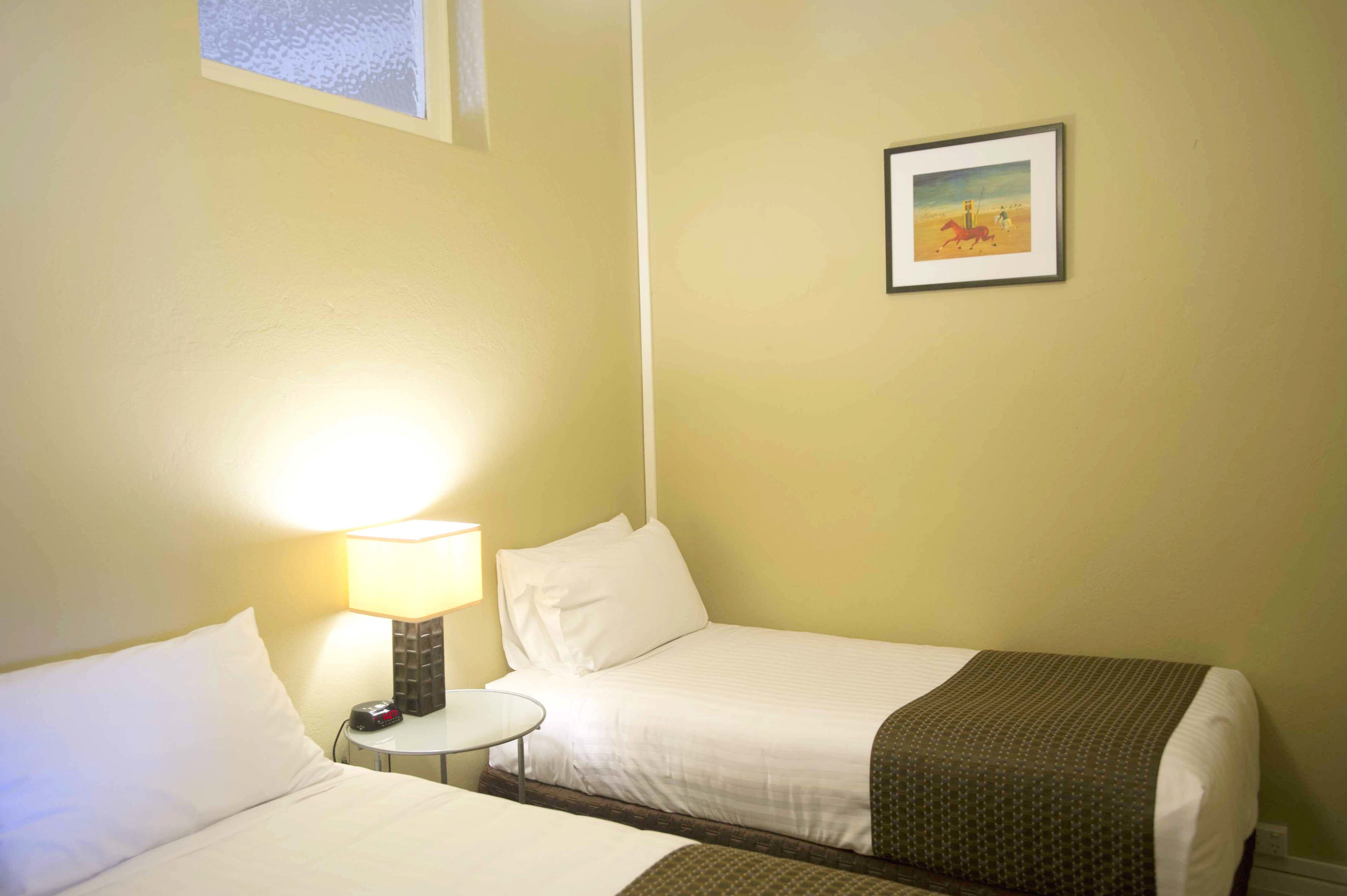 Une chambre d’hôtel avec deux lits, une table de chevet avec une lampe, une petite table et un tableau encadré au mur.