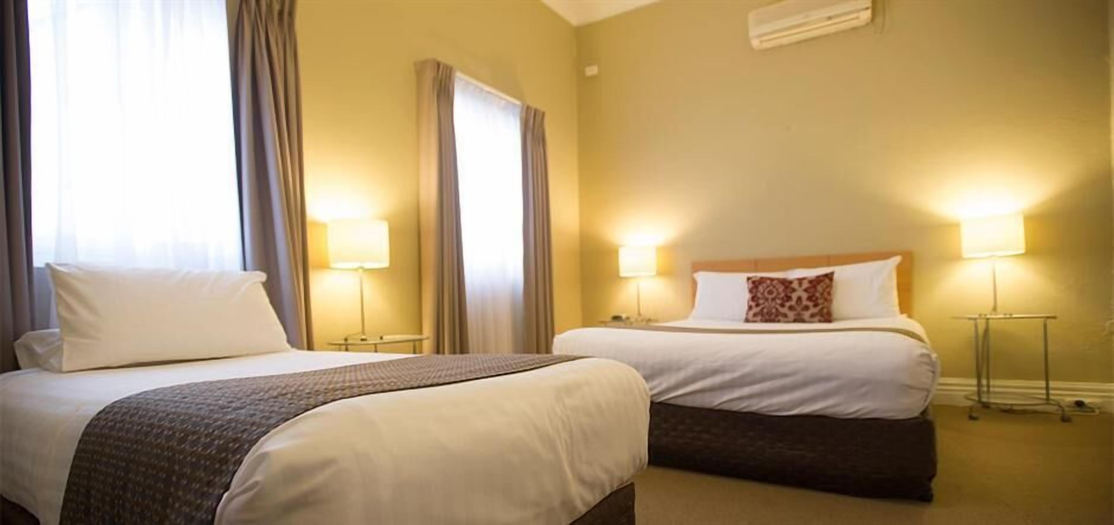 A hotel room with two beds, each with a bedside lamp, a small table, and a window with curtains.