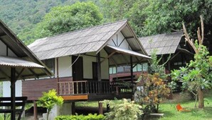 Una casa de madera tradicional con balcón, rodeada de exuberante vegetación y montañas al fondo.