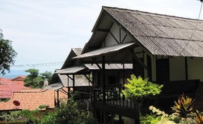 A traditional wooden house with a balcony, surrounded by lush greenery and a view of a red-roofed building in the background.