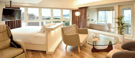 A modern hotel room with a bed, seating area, TV, and large windows.