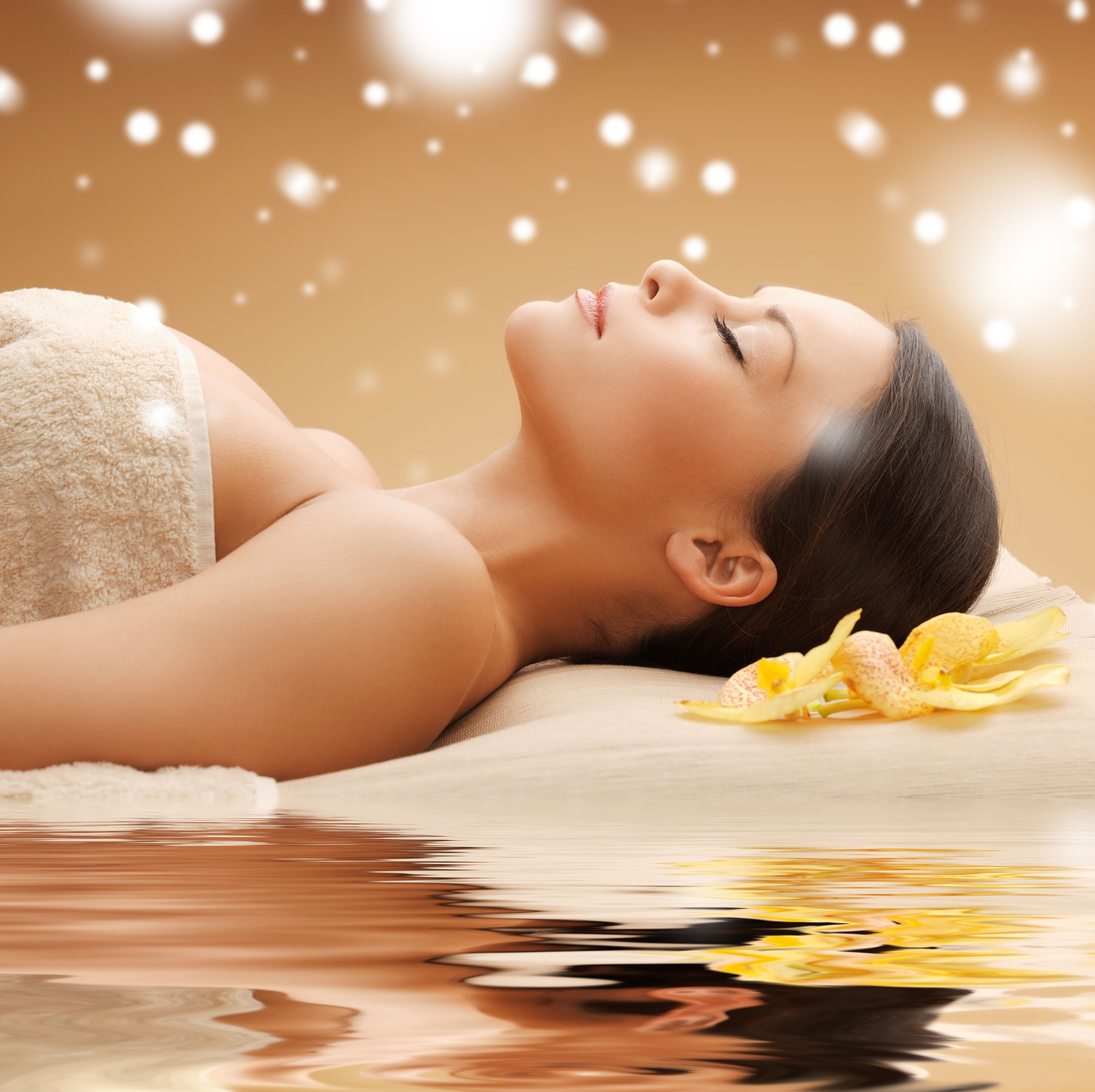 A person lying on a massage table with a towel wrapped around their upper body, a flower beside them, and a reflection on the water surface.