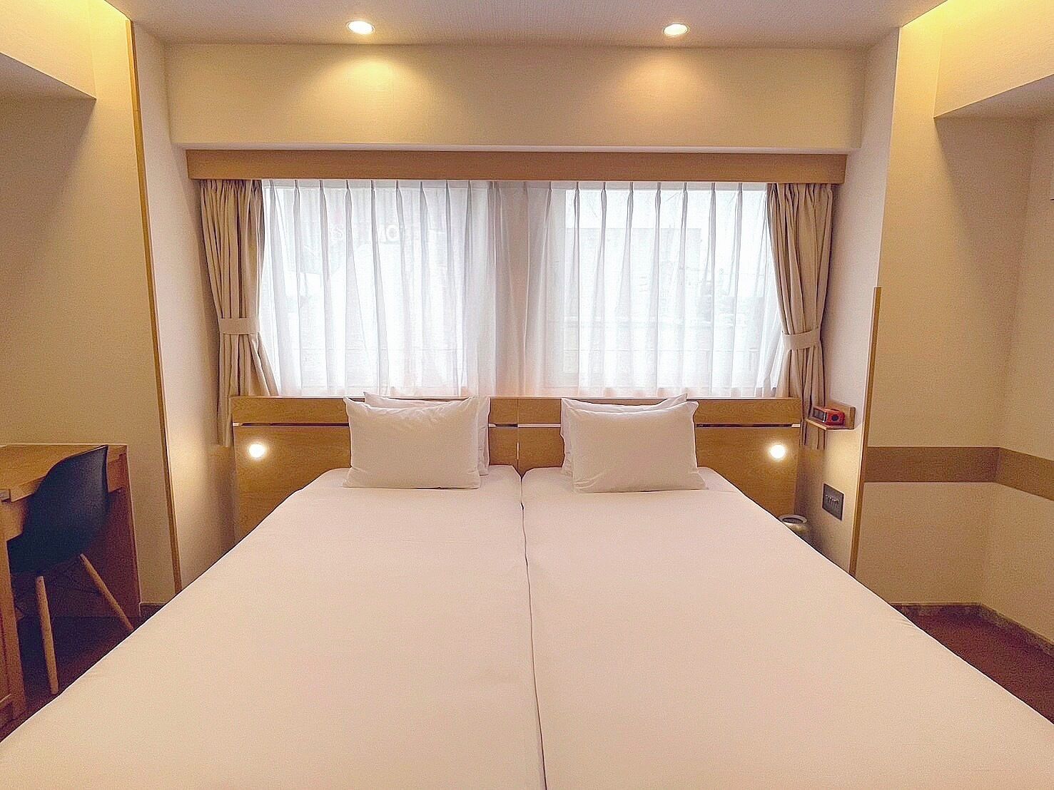 A double bed with white linens, a wooden headboard, and a window with sheer curtains.