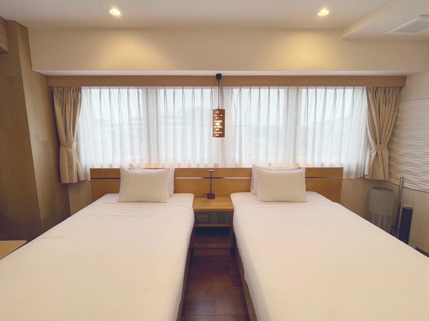 Una camera d'albergo con due letti, un'ampia finestra con tende, un comodino con una lampada e un punto luce a parete.