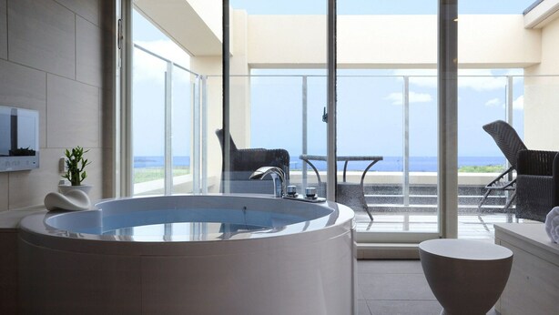A modern bathroom with a large bathtub, a small table, and a chair. There is a large window providing a view of the ocean. - Breath Hotel (Fujisawa)
