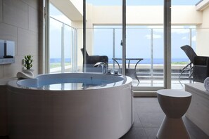 A modern bathroom with a large bathtub, a small table, and a chair. There is a large window providing a view of the ocean. - Breath Hotel (Fujisawa)