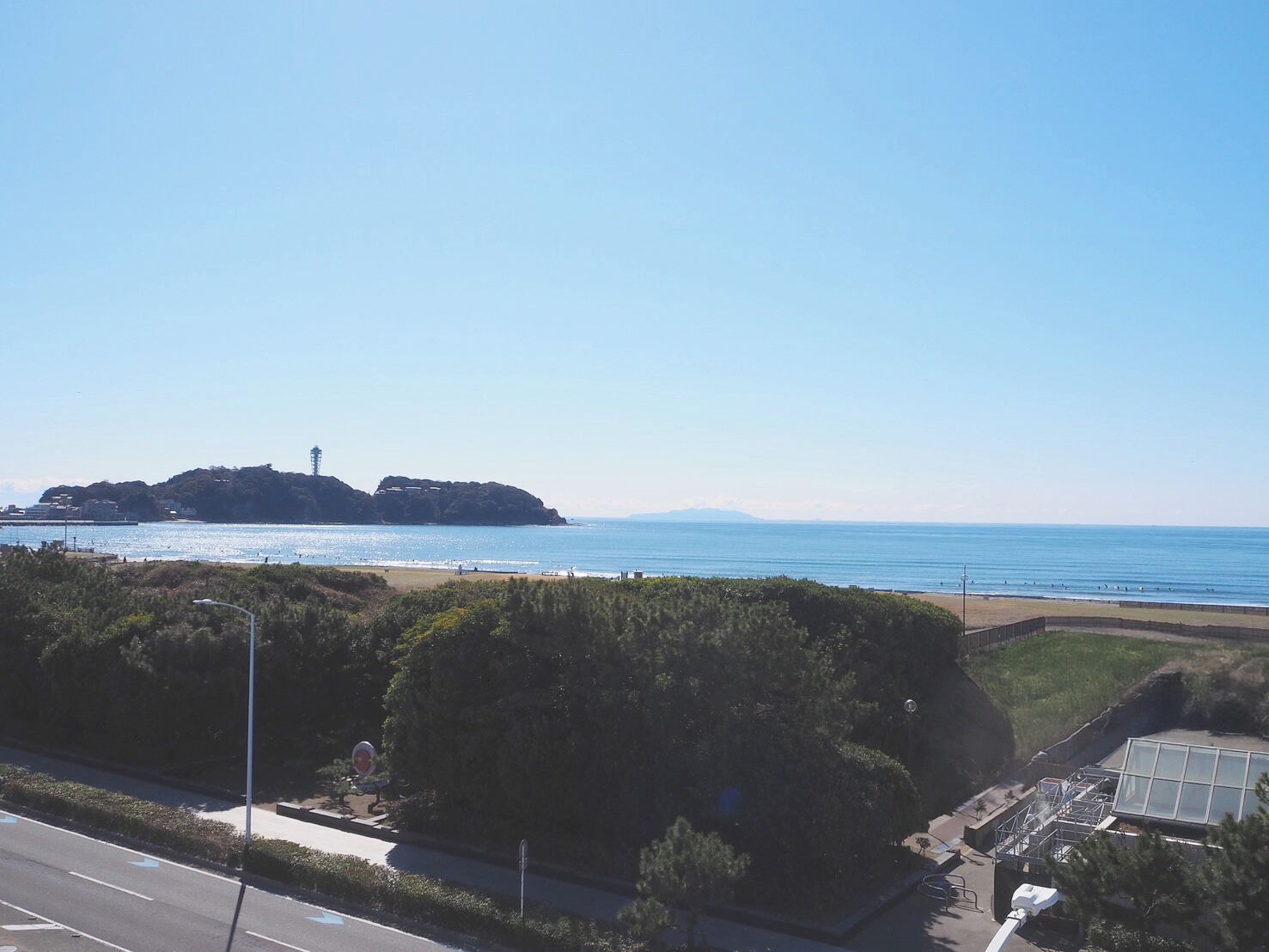 Une vue côtière avec un phare situé sur une île, une plage et une route bordée de verdure au premier plan.