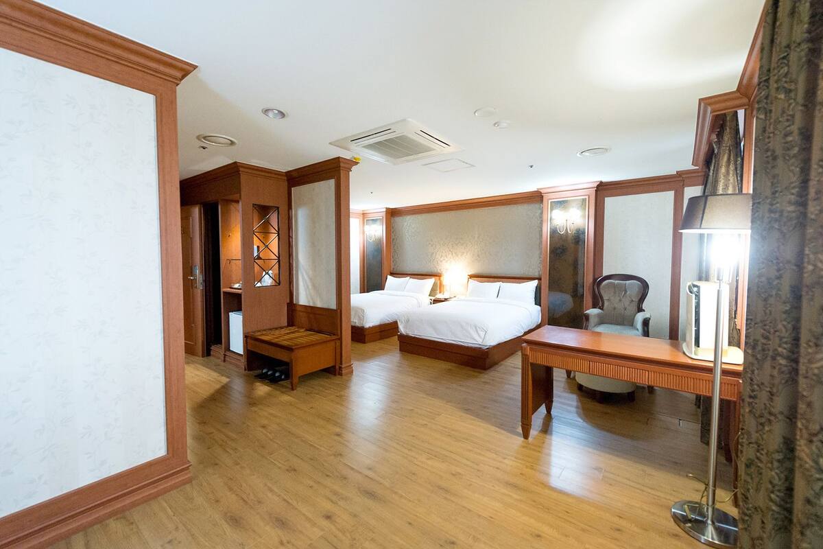 A hotel room with two beds, a wooden floor, a desk, a chair, a mirror, and a wardrobe.
