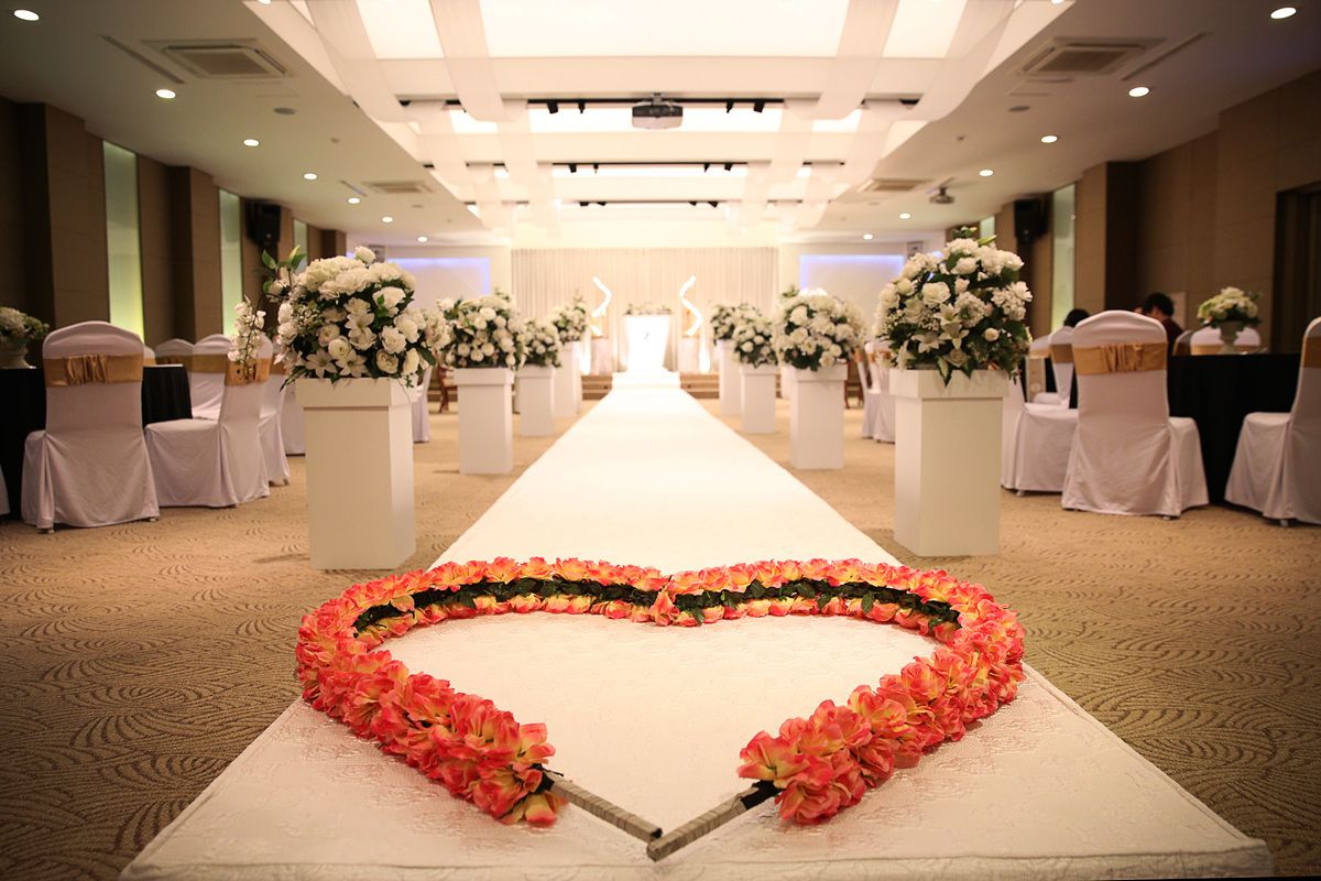A hotel corridor with a white carpeted floor, white chairs with gold sashes, and large white floral arrangements on pedestals.