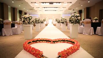 A hotel corridor with a white carpeted floor, white chairs with gold sashes, and large white floral arrangements on pedestals.