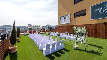 A rooftop wedding setup with rows of white chairs and tables, potted plants, and a view of the cityscape.