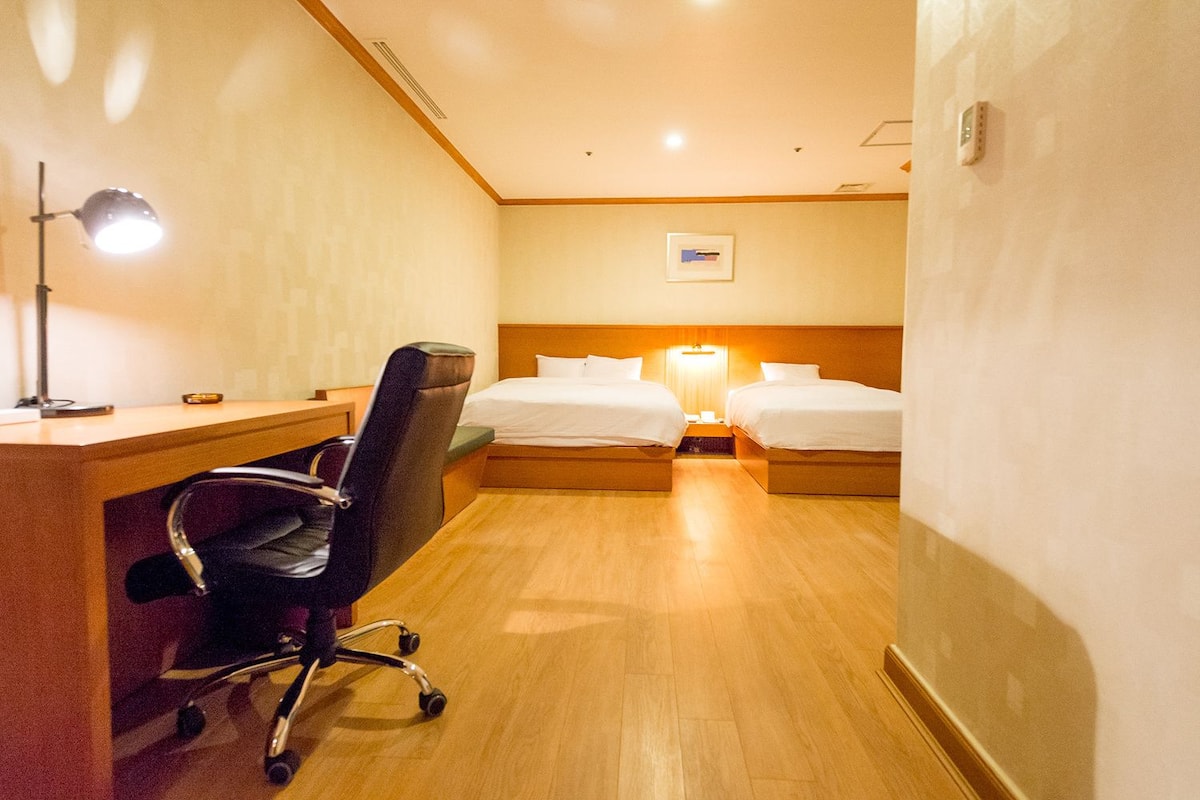 A hotel room with a desk, two single beds, a chair, and a lamp.