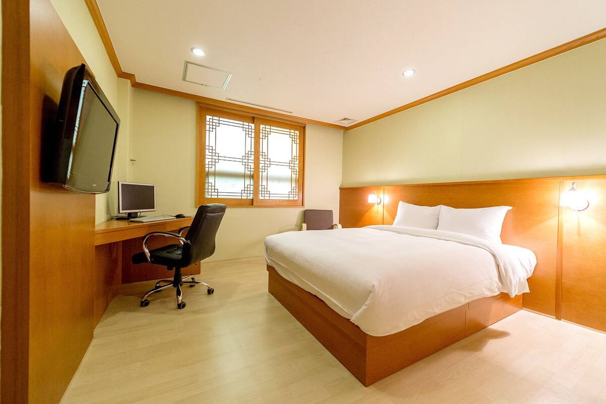 A hotel room with a bed, desk, chair, TV, and window with decorative grille.