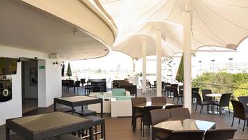 Un área de estar al aire libre con mesas y sillas, cubierta por sombrillas grandes, y una vista del paisaje urbano al fondo.