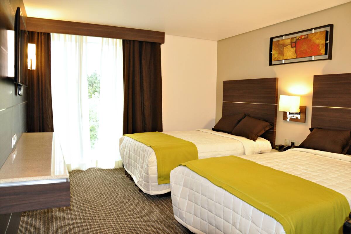 A hotel room with two beds, a flat-screen TV, a painting on the wall, and a window with curtains.