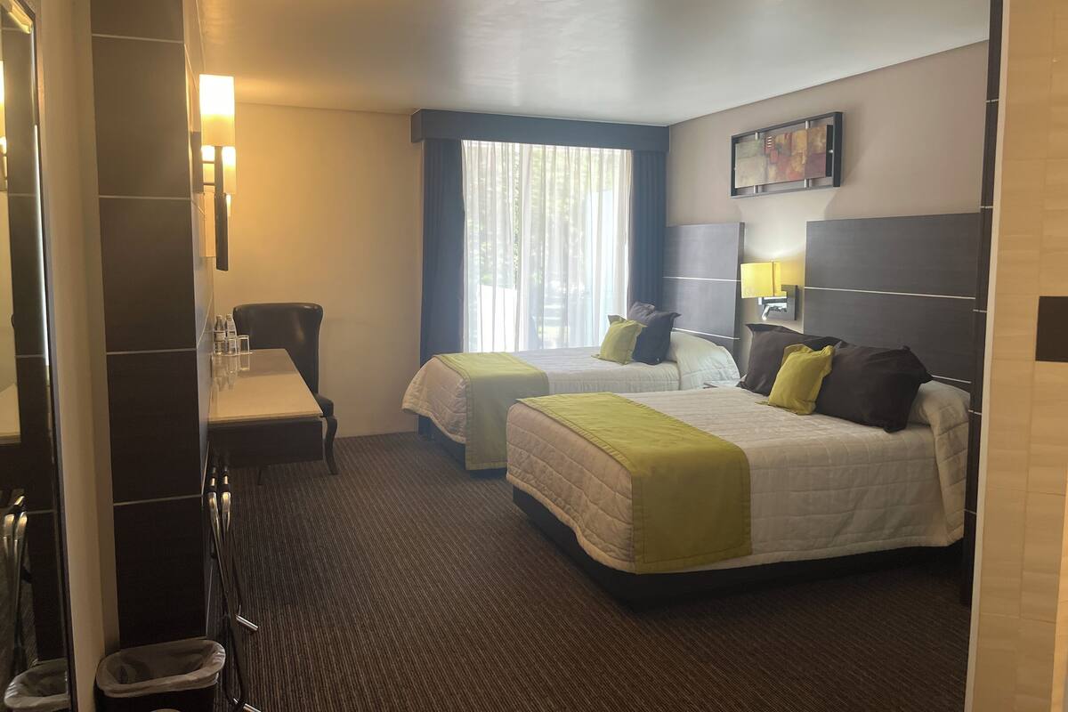 A hotel room with two beds, a desk, a chair, a small trash bin, and a window with curtains.
