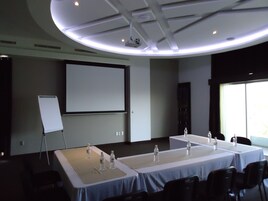 Un salón de conferencias con dos mesas, sillas, una pantalla de proyector y una pizarra.