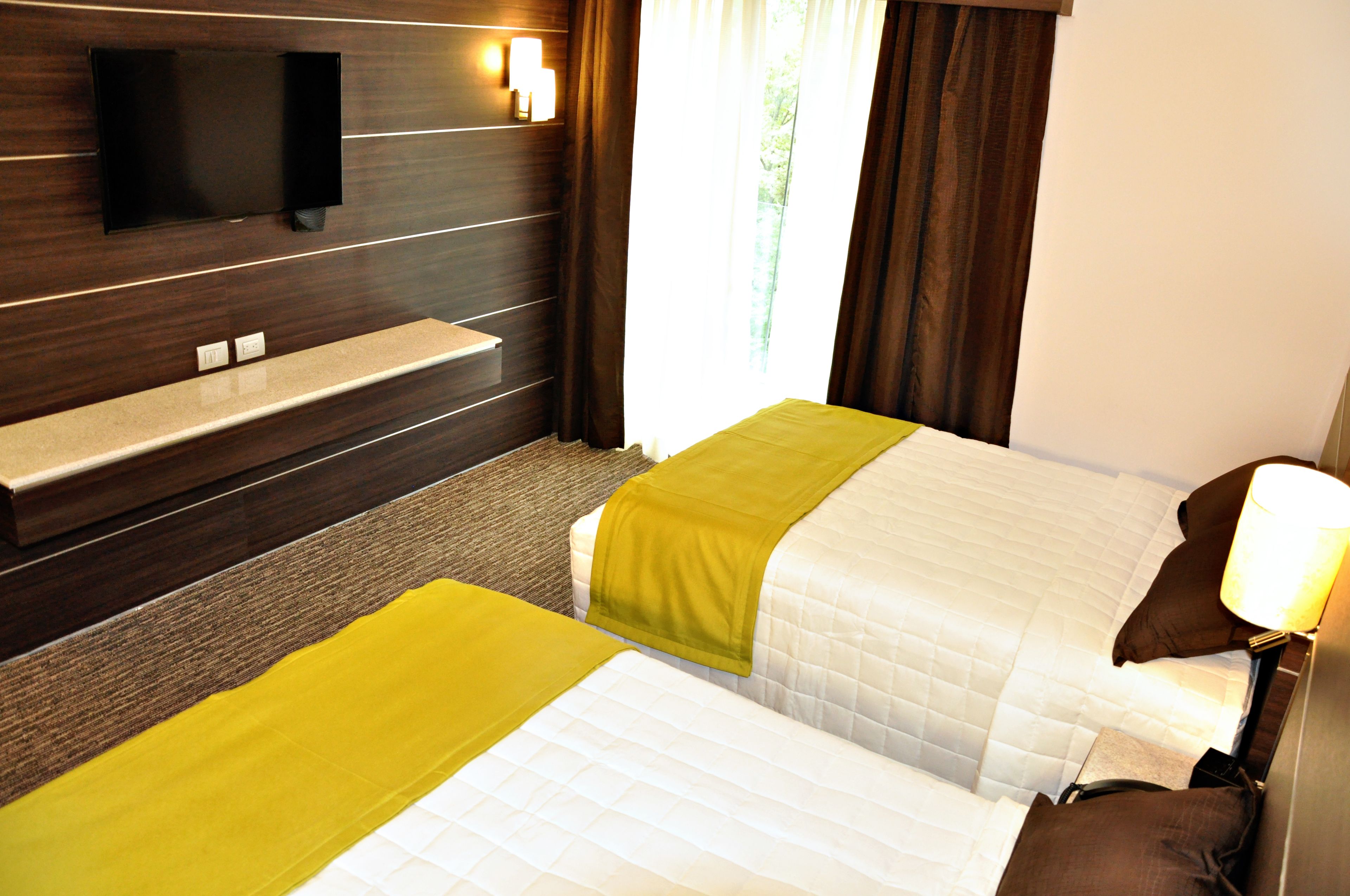 Een hotelkamer met twee bedden, een aan de muur bevestigde televisie, een bankje en een raam met gordijnen.