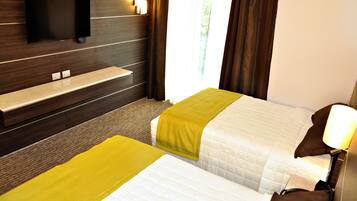 Een hotelkamer met twee bedden, een aan de muur bevestigde televisie, een bankje en een raam met gordijnen.