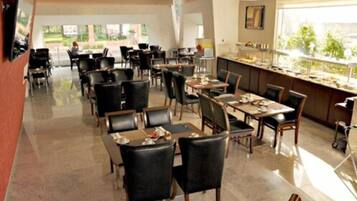 Un comedor de hotel con varias mesas y sillas, una zona de cocina y ventanas amplias que ofrecen vistas al exterior.