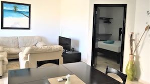 A modern living room with a beige sofa, a dining table with a remote control and a vase, and a framed picture of a beach scene. - La Playa Condo (Playa del Carmen)