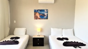 A hotel room with two beds, a nightstand with a lamp, an air conditioner, and a painting of a mountain landscape. - La Playa Condo (Playa del Carmen)