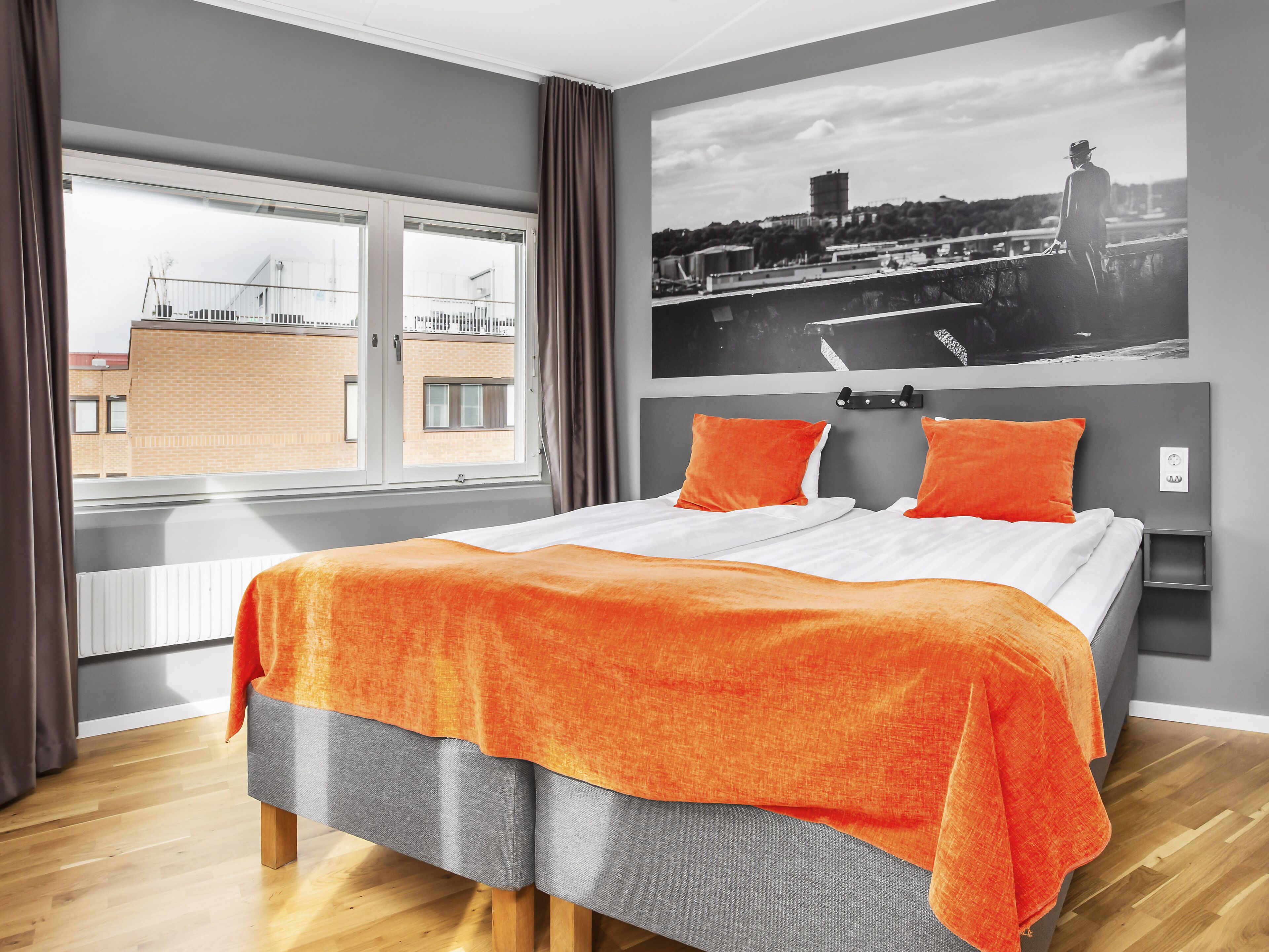 A bedroom with a large bed, grey and orange bedding, a cityscape photo on the wall, and a window with curtains.
