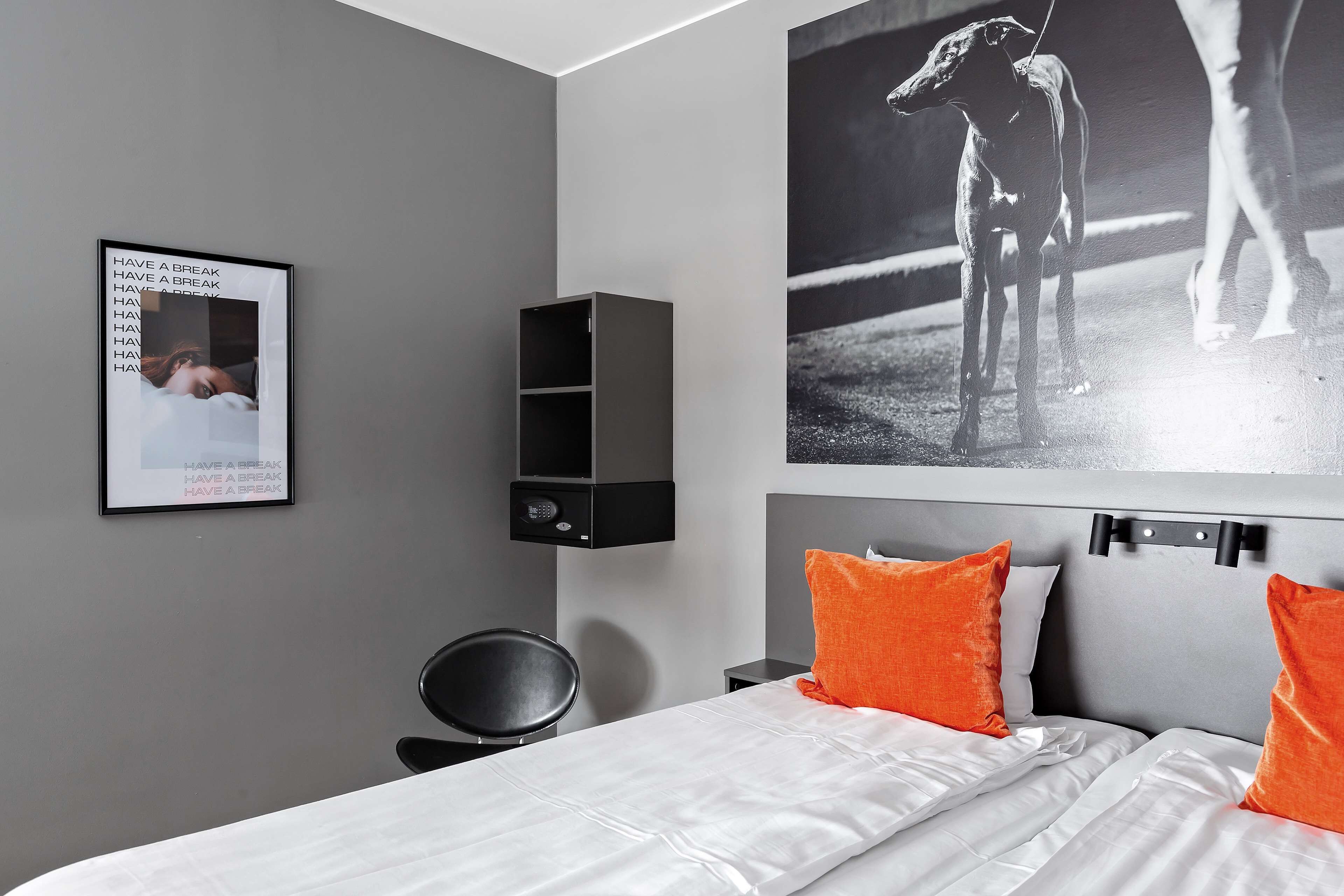 Una habitación de hotel moderna con una pared gris, un estante negro, una cama con almohadas blancas y anaranjadas, y un cuadro enmarcado en la pared.