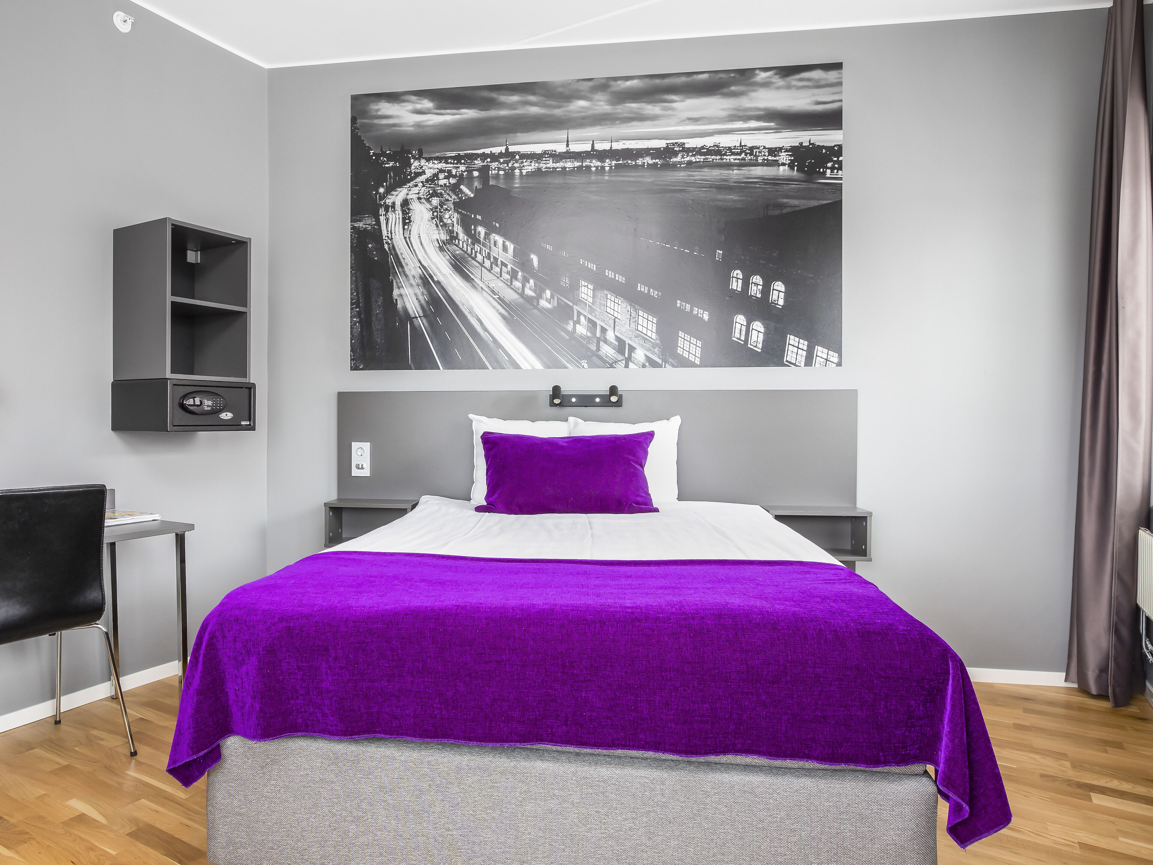 Een hotelkamer met een bed, nachtkastje, stoel, plank en een groot stadsgezicht op de muur.