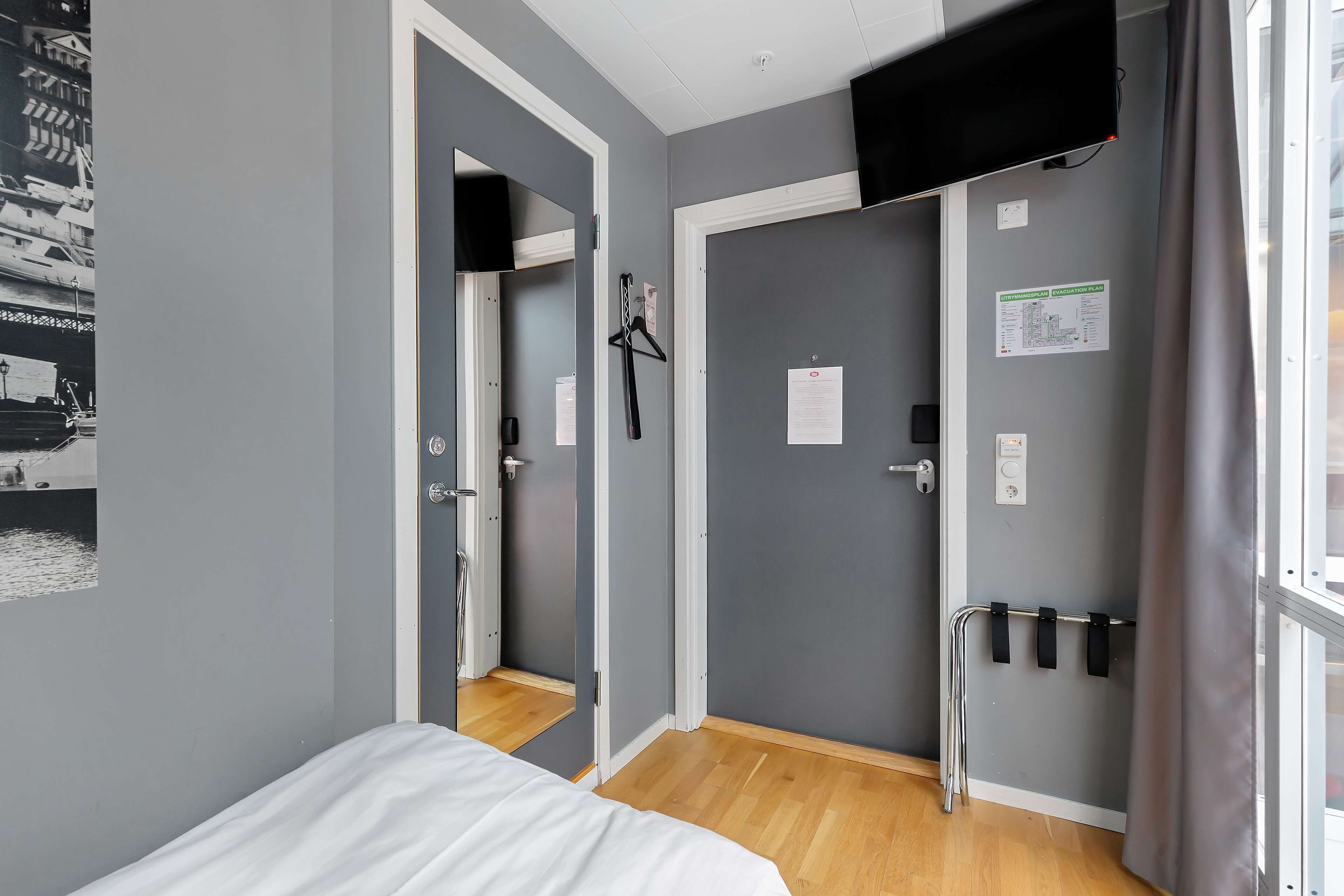 A hotel room with a bed, a mirror, a door with a coat rack, and a window with curtains.