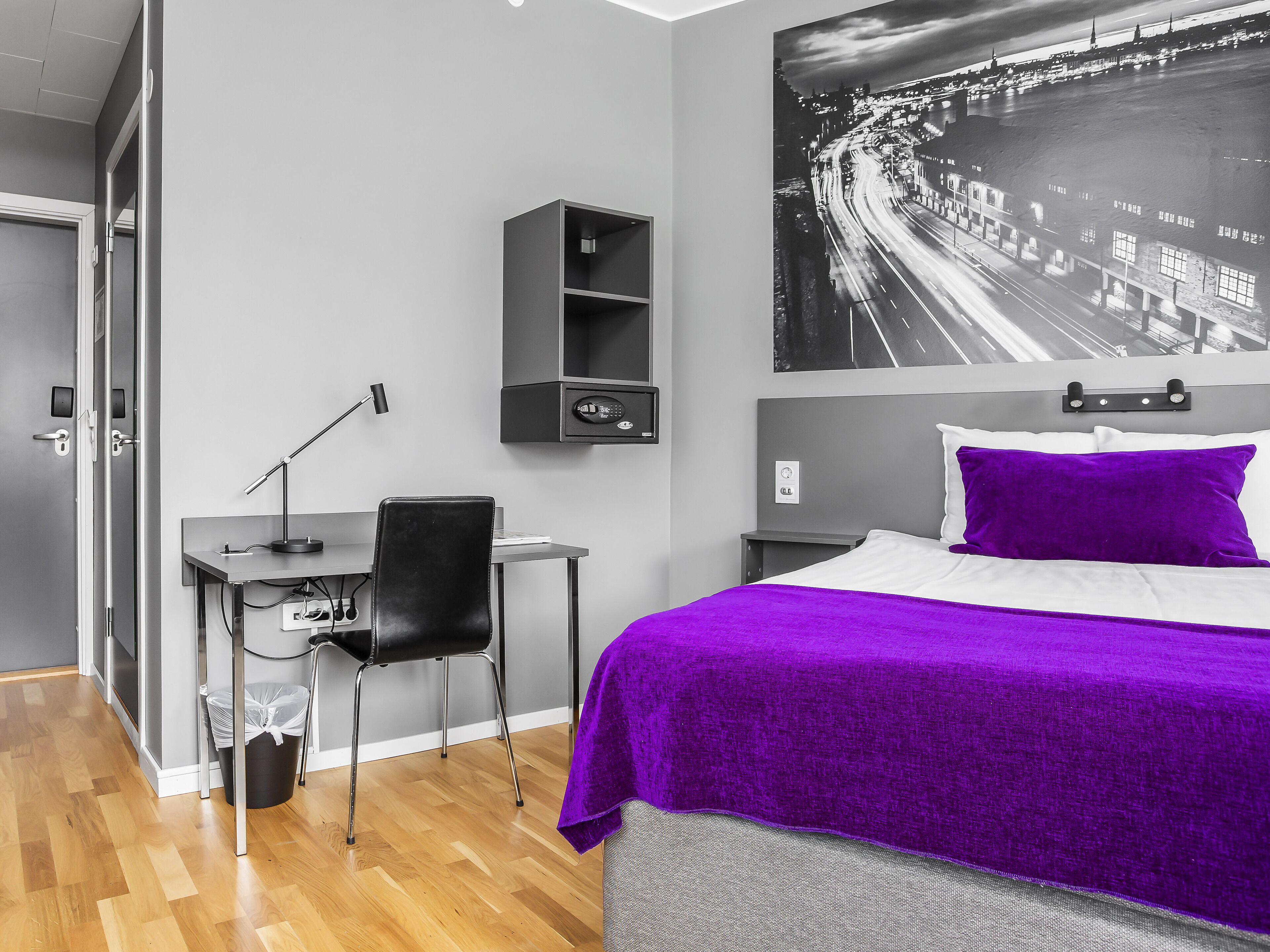 A modern hotel room with a bed, a desk, and a chair. There is a large window with a view of a cityscape.