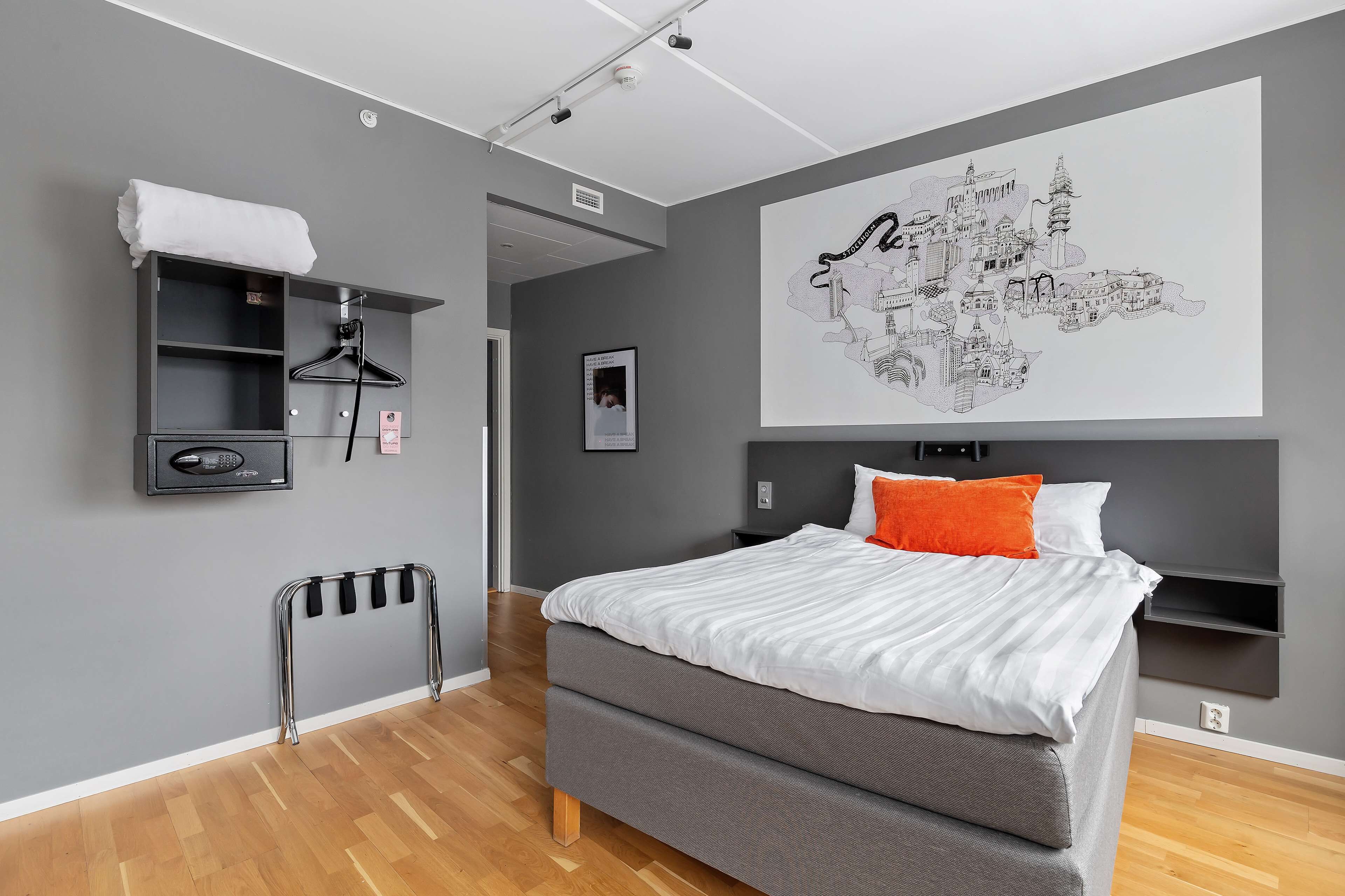 A modern bedroom with a grey bed, white bedding, a towel on a shelf, a hanging rack, and a cityscape artwork on the wall.