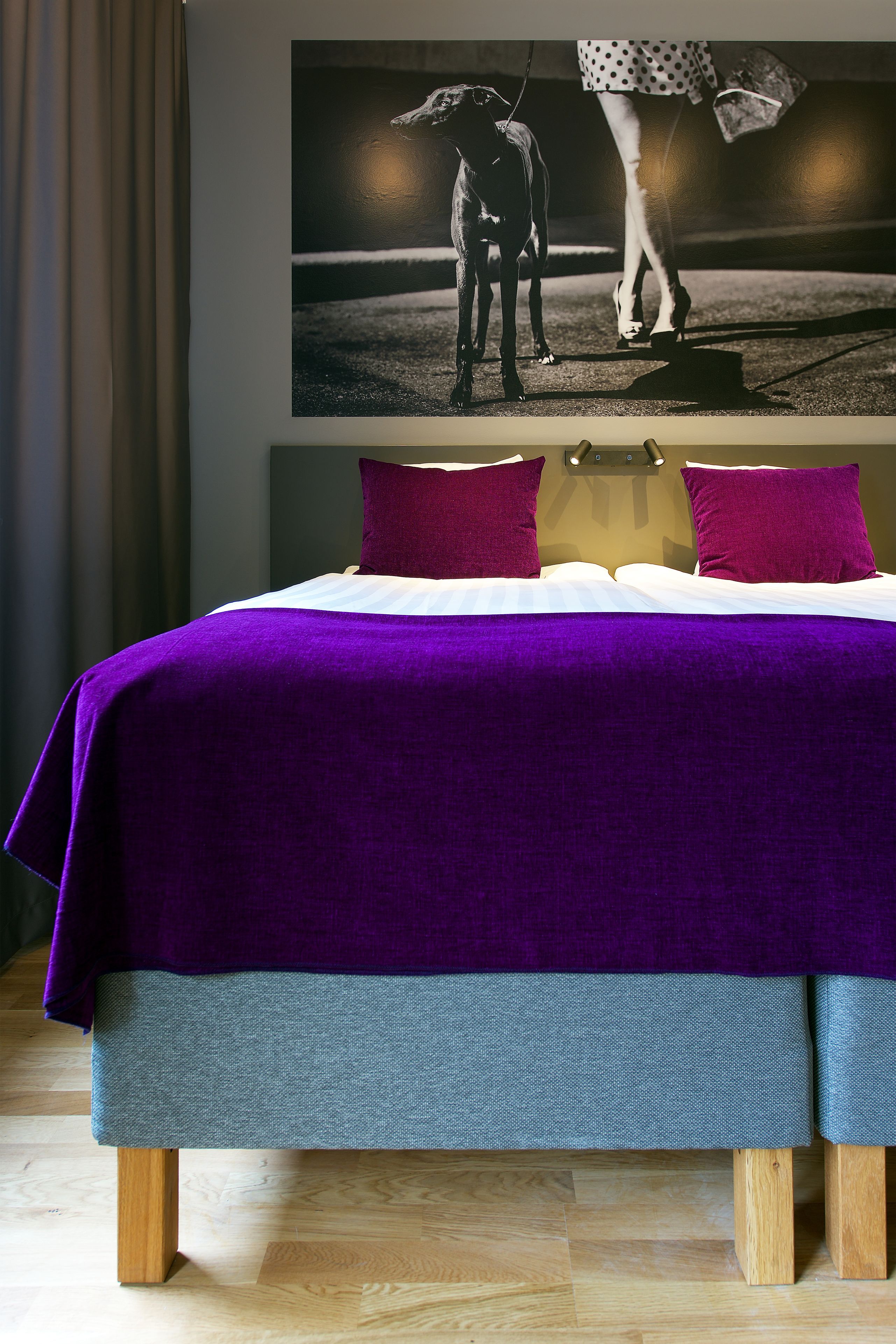 Una cama con colcha morada y gris, patas de madera y dos almohadas.