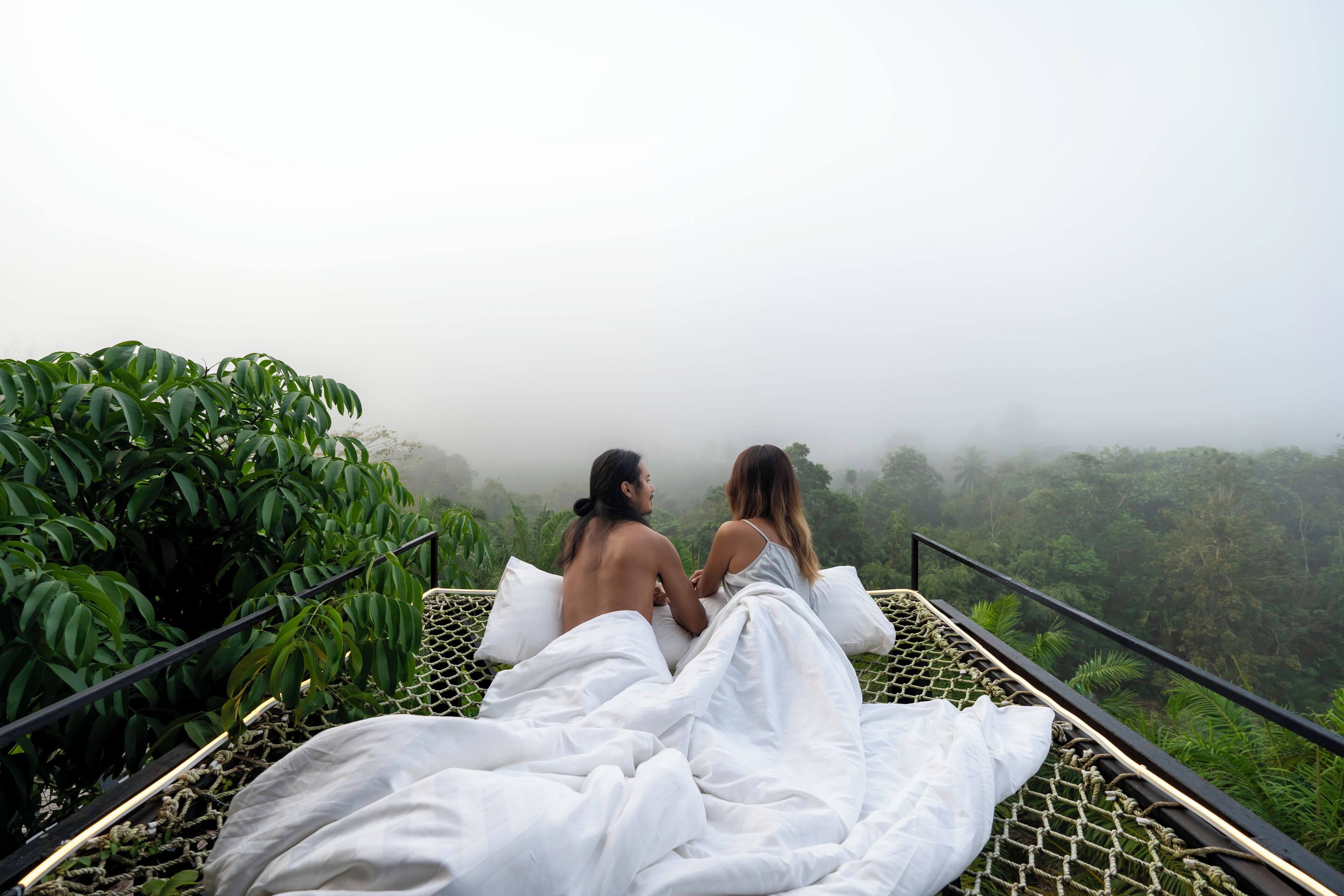 Two individuals sitting on a bed with white sheets, overlooking a lush forested landscape.