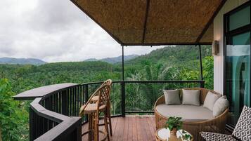 Un balcón con mobiliario de mimbre, un rincón de estar con cojines y vistas a vegetación exuberante y montañas.