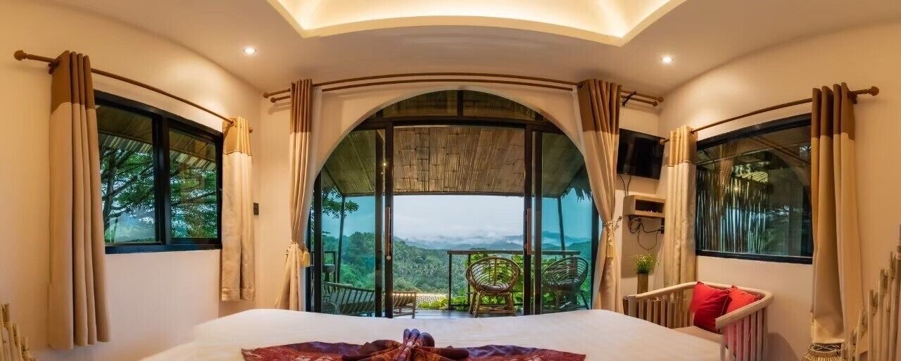 A bedroom with a large bed, a view of the outdoors, and a balcony with chairs.