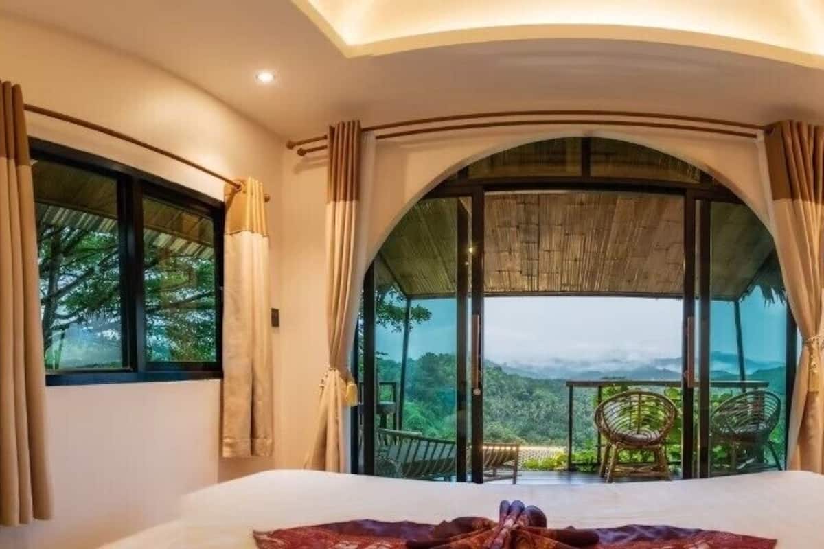 A bedroom with a large bed, a view of the outdoors, and a balcony with chairs.