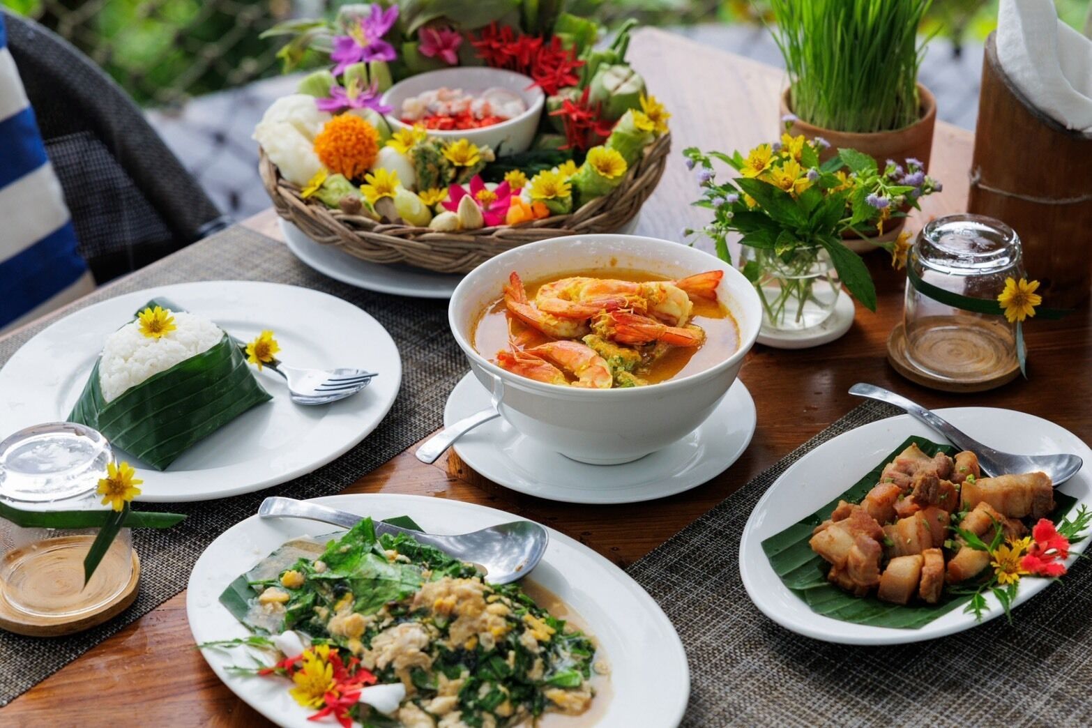 Mesa dispuesta con diversos platos, incluyendo arroz, sopa y carne, rodeada de flores y una cesta de frutas.